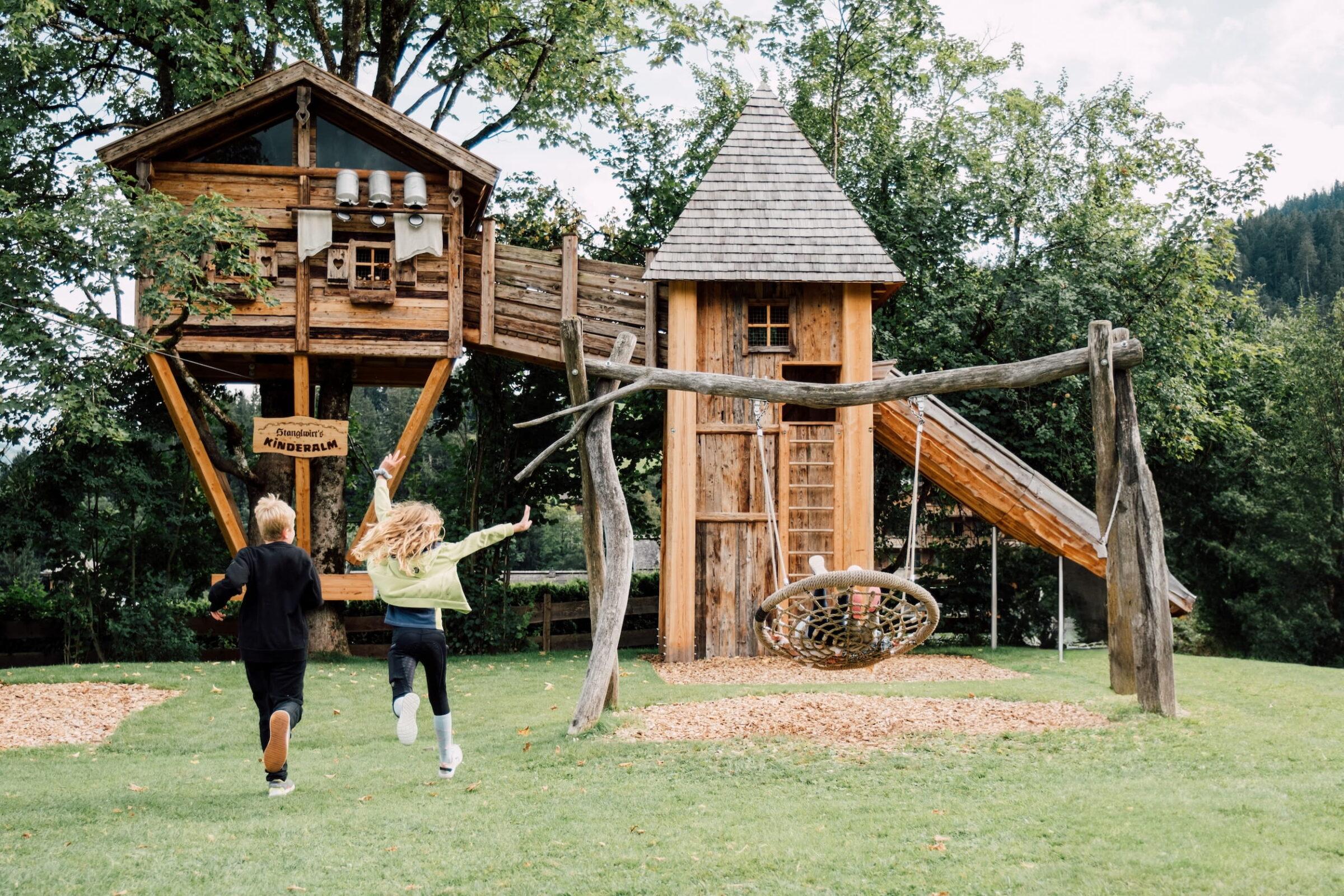 Kinder rennen auf eine aus Holz gebaute Spielanlage mit Baumhaus, Rutsche und Nestschaukel im Bio- und Wellnessresort Stanglwirt in Tirol