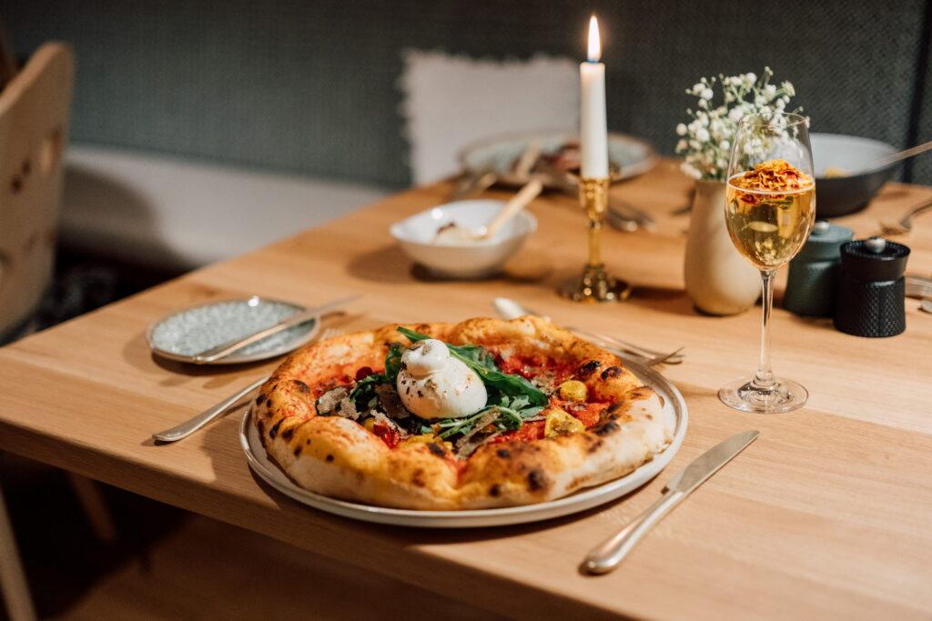 Pizza mit Burrata, Spinat und Tomaten auf einem Teller auf einem Holztisch mit Kerze, Weinglas und Blumenvasen im Hintergrund