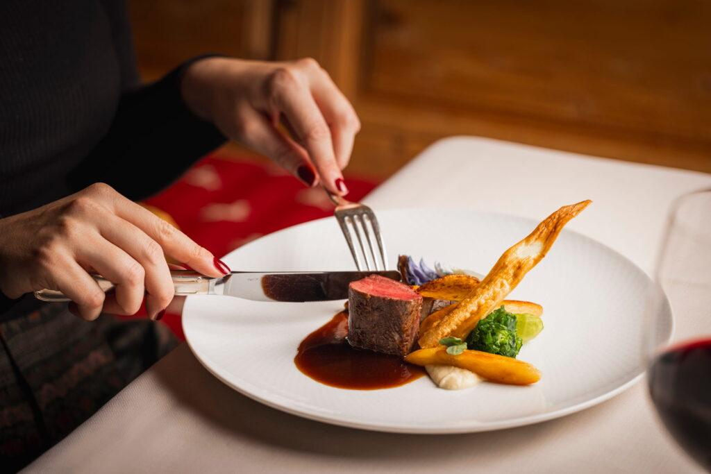 Person schneidet ein kunstvoll angerichtetes Gericht mit Fleisch, Gemüse und Sauce auf einem weißen Teller im Bio- und Wellnessresort Stanglwirt in Tirol