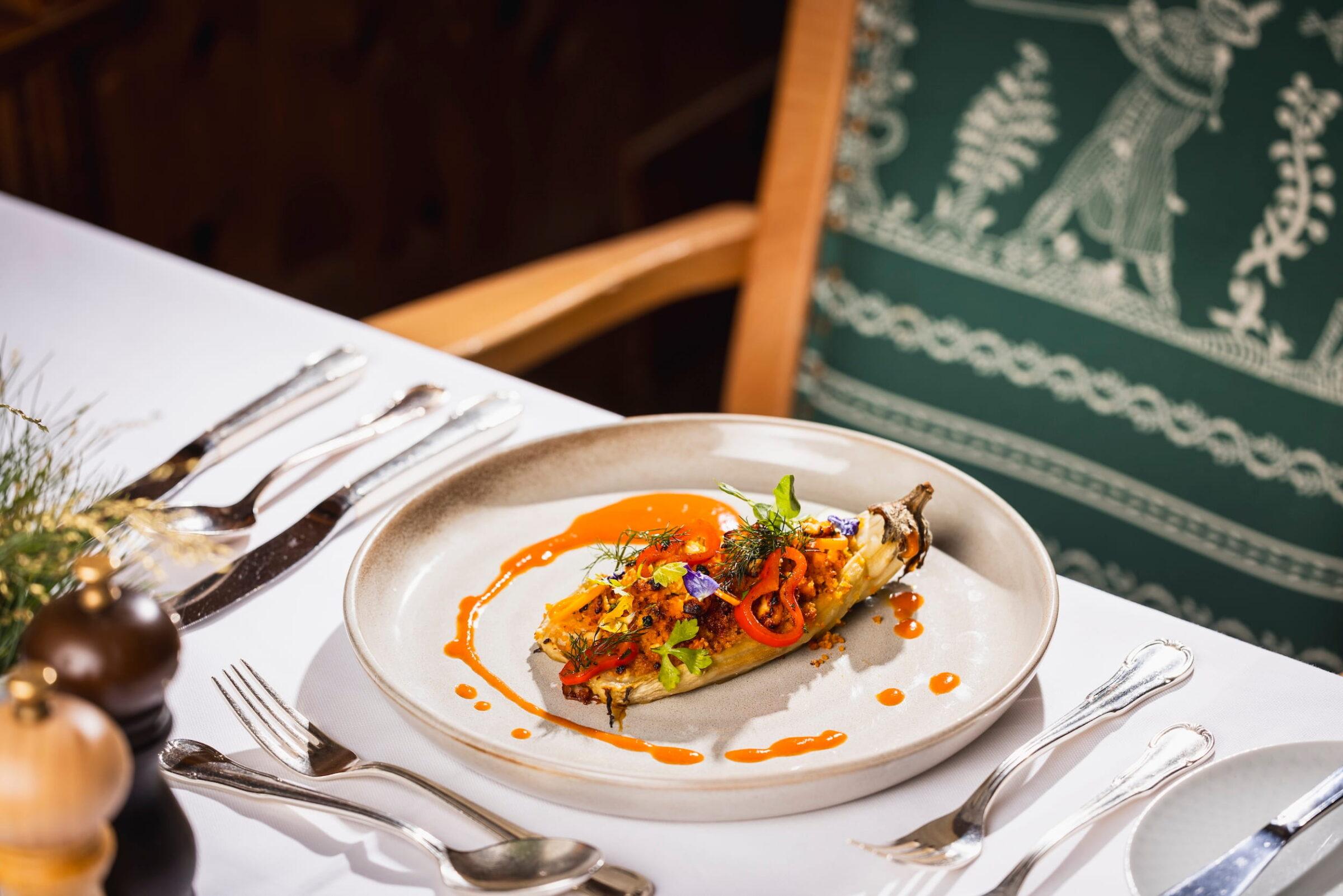 Auf einem weißen Teller liegt ein vegetarisches Gericht mit gegrillter Aubergine, roten Paprikastreifen, Kräutern und oranger Sauce, angerichtet auf einem weiß gedeckten Tisch mit Besteck und grün gemustertem Stuhl im Hintergrund | in einem Restaurant des Stanglwirts in Tirol