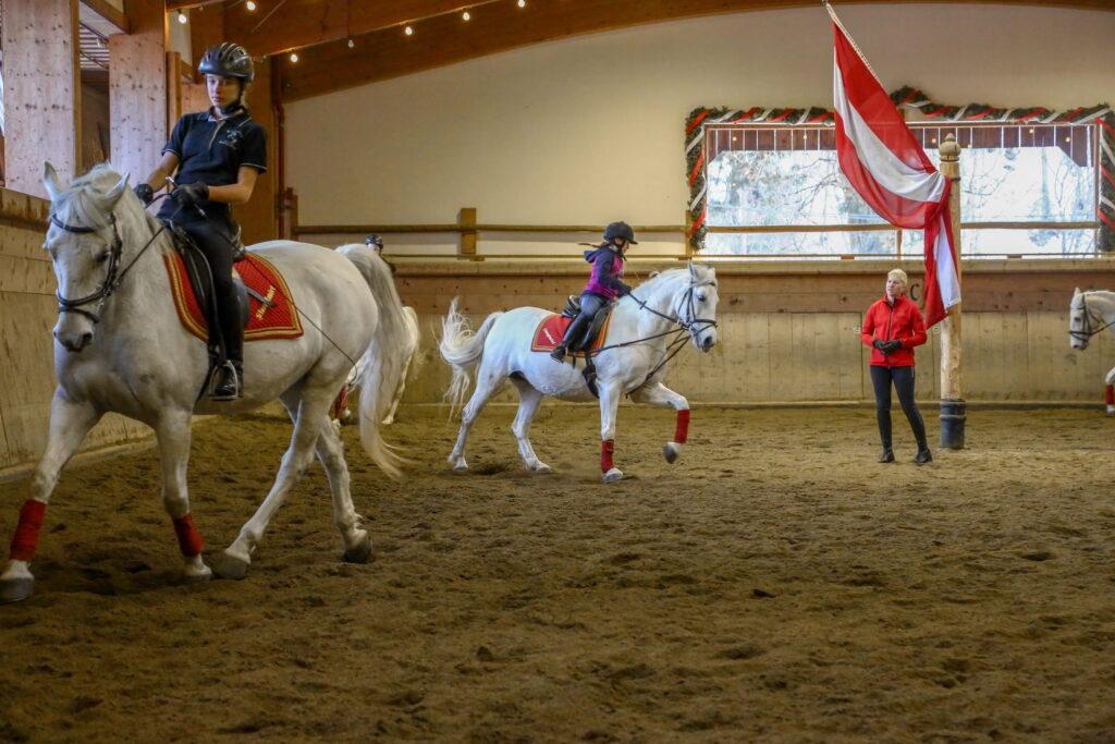 Reitstunde in der Reithalle des Bio- und Wellnessresorts Stanglwirt mit mehreren Reitern auf weißen Pferden und einer stehenden Person neben einer Fahnenstange mit rot-weiß-roter Fahne