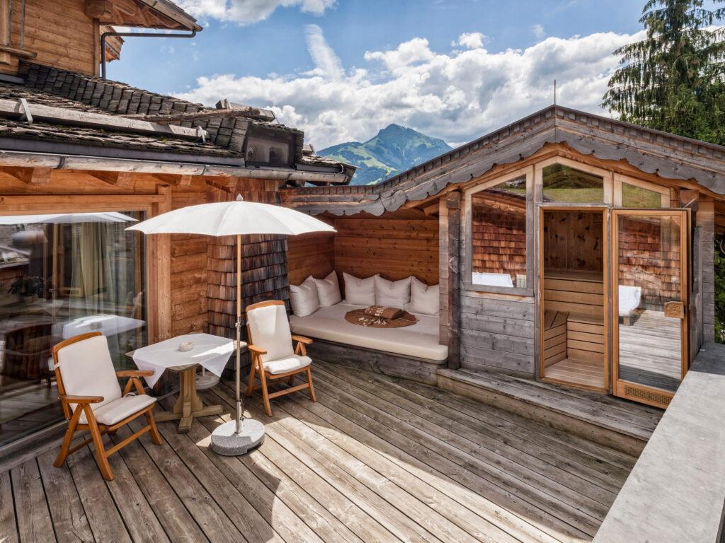 Eine rustikale Holzterrasse mit gemütlichen Sitzgelegenheiten, einem weißen Sonnenschirm und einem kleinen Tisch unter strahlend blauem Himmel im Bio- und Wellnessresort Stanglwirt in Tirol. Berge und Kiefern bilden eine ruhige Kulisse.