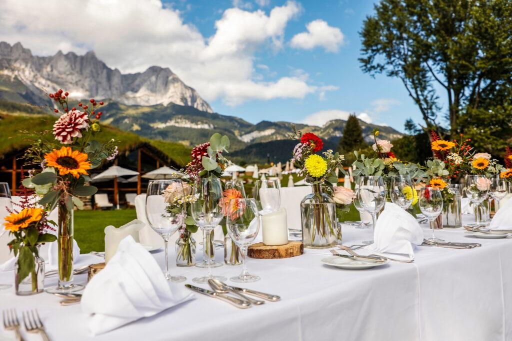 Festlich gedeckter Tisch mit weißen Tischdecken, gefalteten Servietten, Gläsern, Besteck und bunten Blumenarrangements vor Bergpanorama im Bio- und Wellnessresort Stanglwirt in Tirol