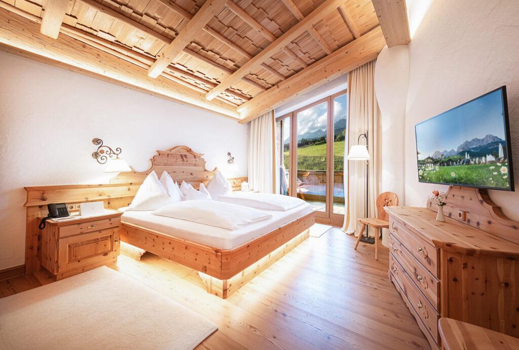 Ein modernes Schlafzimmer im Landhausstil im Bio- und Wellnessresort Stanglwirt am Wilden Kaiser mit Holzmöbeln, einem großen Bett und einem malerischen Ausblick durch die Glastüren. Die warme Beleuchtung sorgt für eine gemütliche, einladende Atmosphäre.