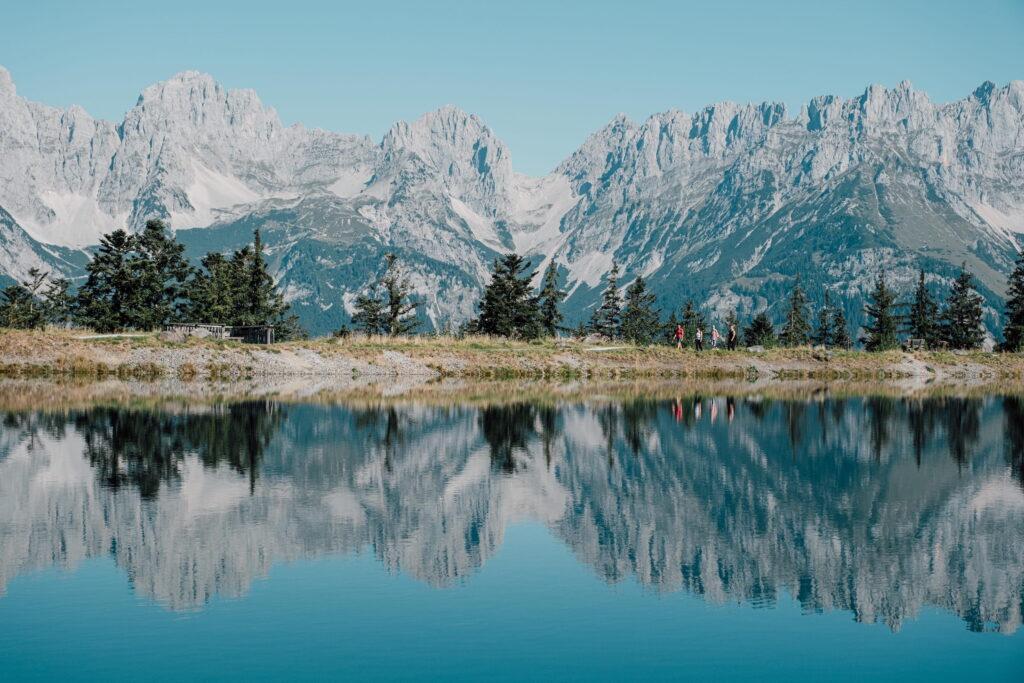 Bergsee mit Spiegelung der Berge und Bäume am Ufer sowie Wanderern am Weg vor den Gipfeln des Wilden Kaisers in Tirol