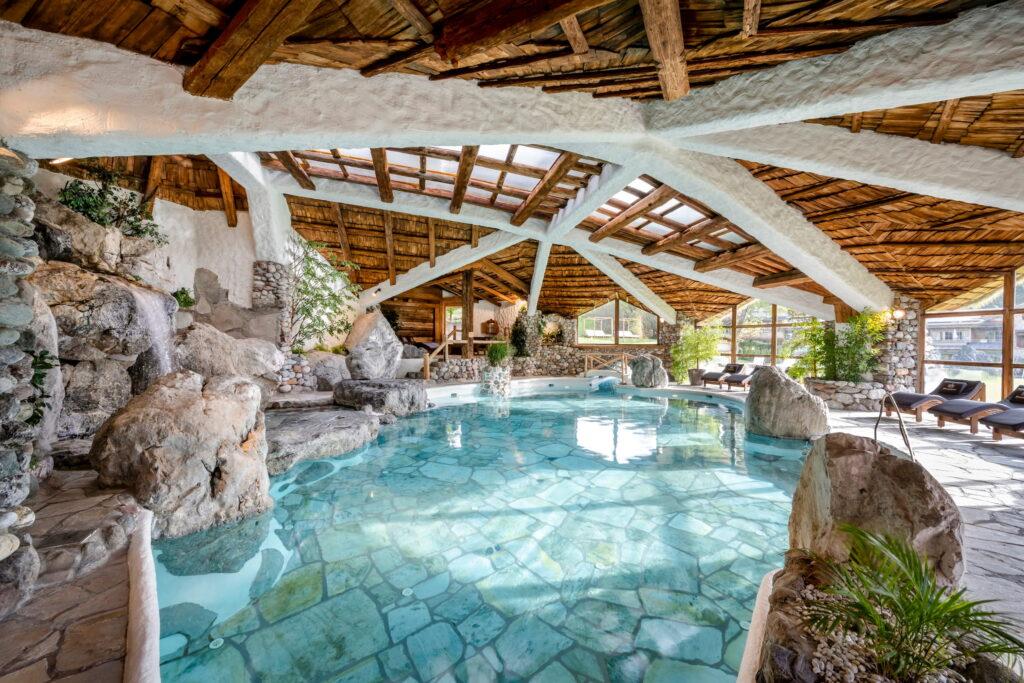 Innenansicht des Felsenbads im Bio- und Wellnessresort Stanglwirt mit natürlicher Steinarchitektur, Wasserfall, Holzdecke und Liegen am Beckenrand