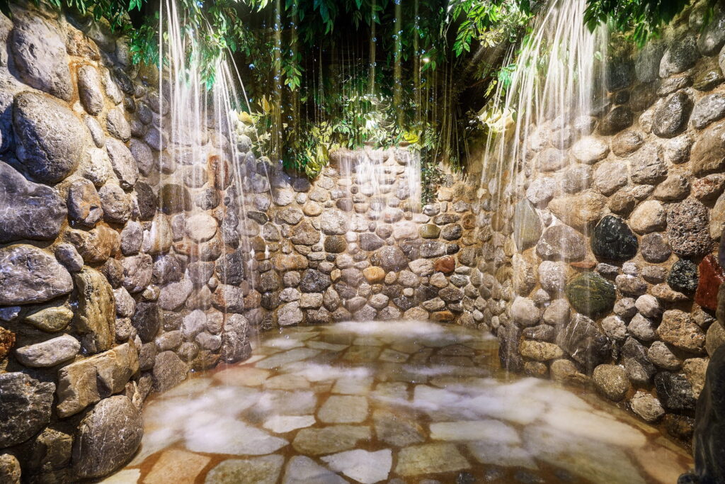 Ein Raum aus Stein im Felsenbad des Bio- und Wellnessresorts Stanglwirt in Tirol, in dem Wasserfälle über die Wände aus großen, glatten Felsen hinabstürzen, umgeben von üppigem Grün. Die Szenerie wirkt ruhig und einladend.