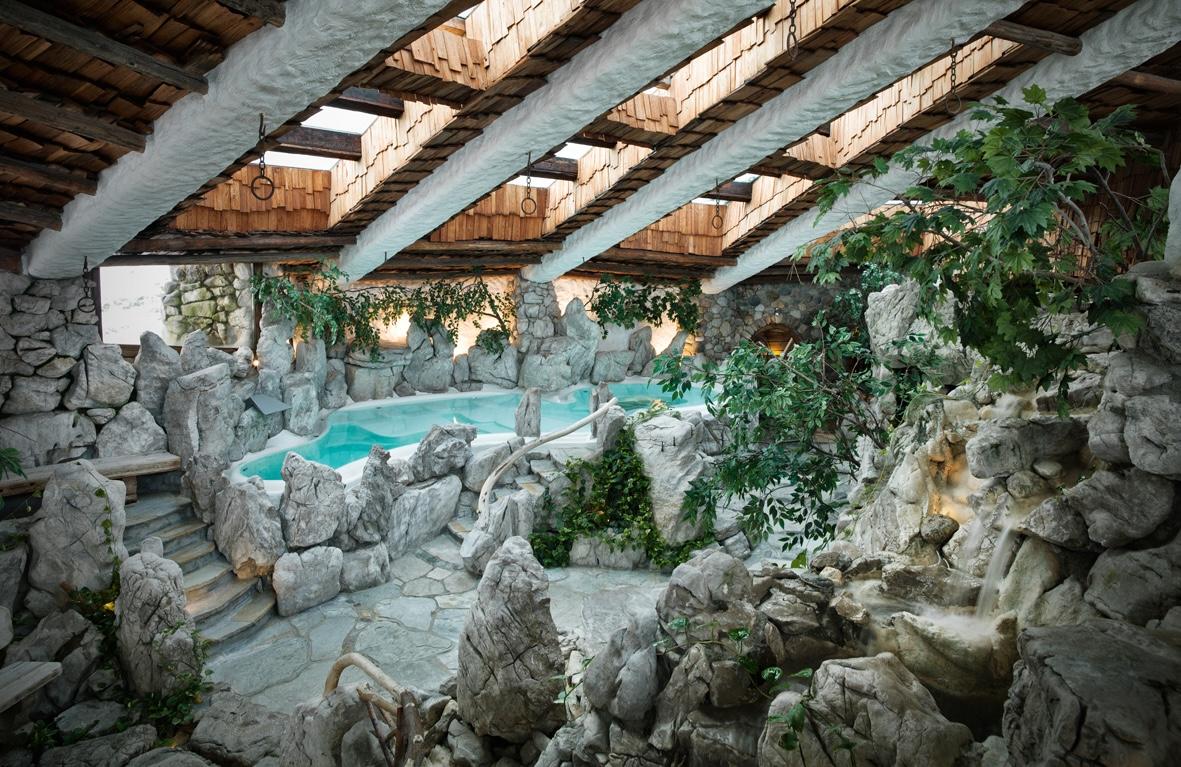 Innenbereich des Wellnessresorts Stanglwirts in Tirol mit natürlicher Felsenpoollandschaft, Wasserfall, Pflanzen und Holzbalkendecke mit Oberlichtern