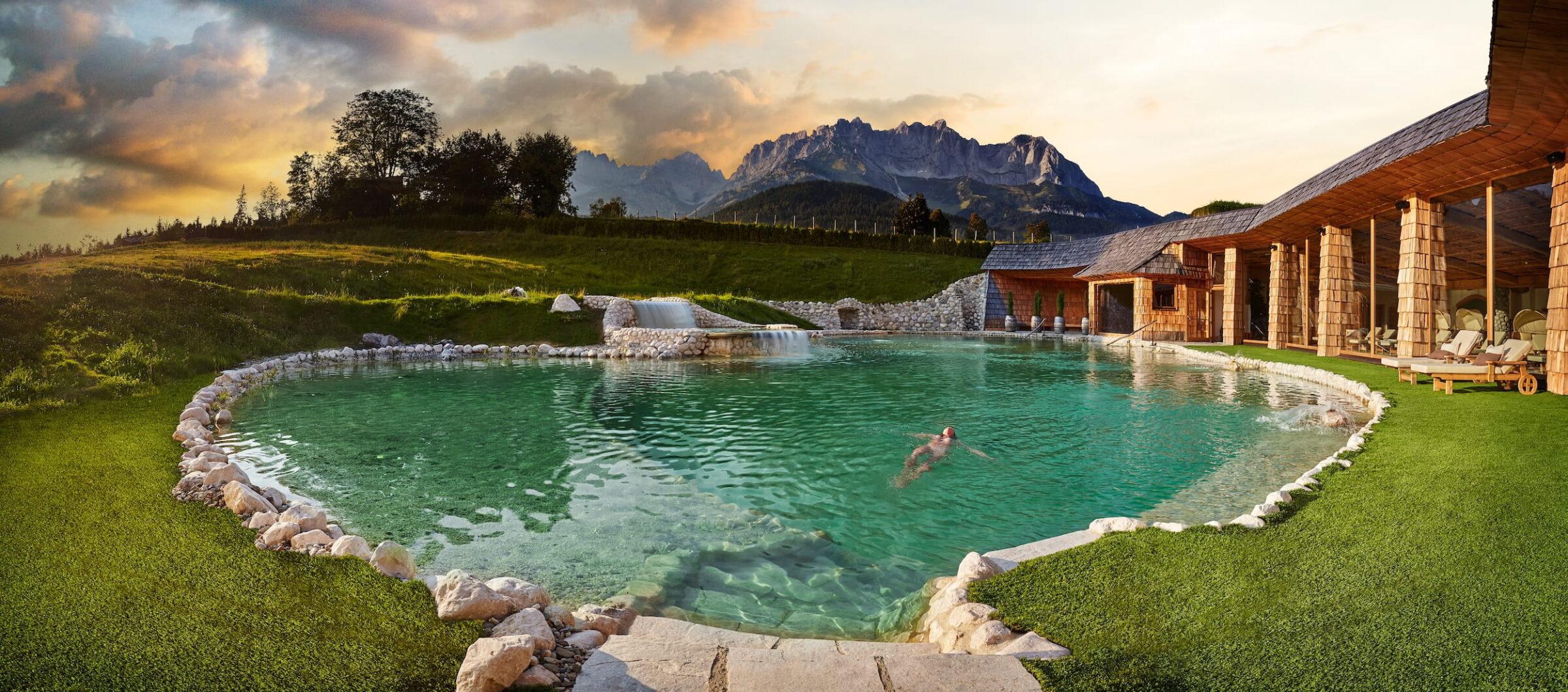 Natürlicher Badesee im Wellnessresort Stanglwirt in Going mit steiniger Umrandung und einer Person im Wasser vor einem Holzgebäude mit Liegestühlen und Bergpanorama des Wilden Kaisers im Hintergrund