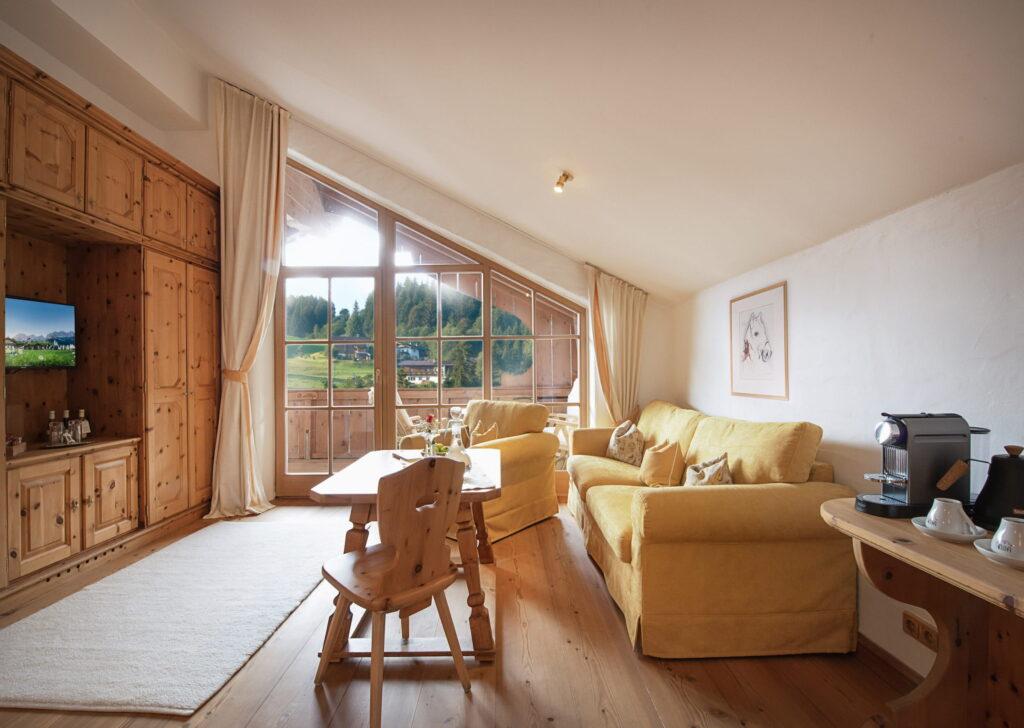 Gemütliches Wohnzimmer im österreichischen Bio- und Wellnessresort Stanglwirt mit Holzböden und warmer Beleuchtung. Gelbe Sofas stehen einem Holztisch gegenüber, und durch die großen Fenster bietet sich ein ruhiger Blick auf den Wald.