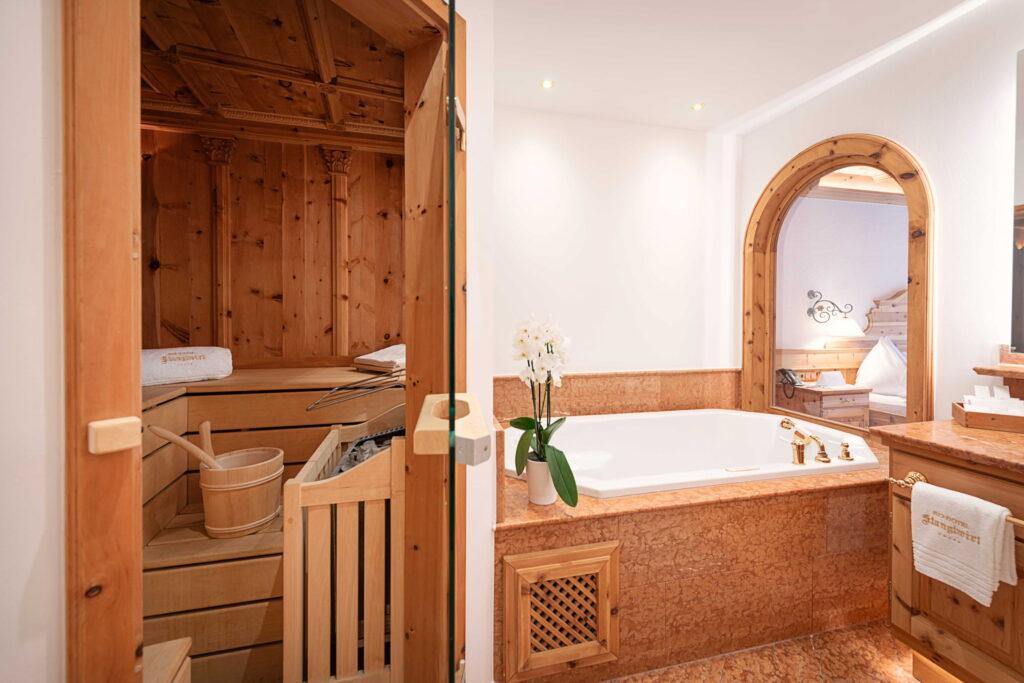 Ein luxuriöses Wellness-Badezimmer in einer Suite im Bio- und Wellnessresort Stanglwirt am Wilden Kaiser mit einer Holzsauna, einer eleganten Marmorbadewanne und sanfter Beleuchtung. Eine Vase mit weißen Orchideen sorgt für eine beruhigende Note.