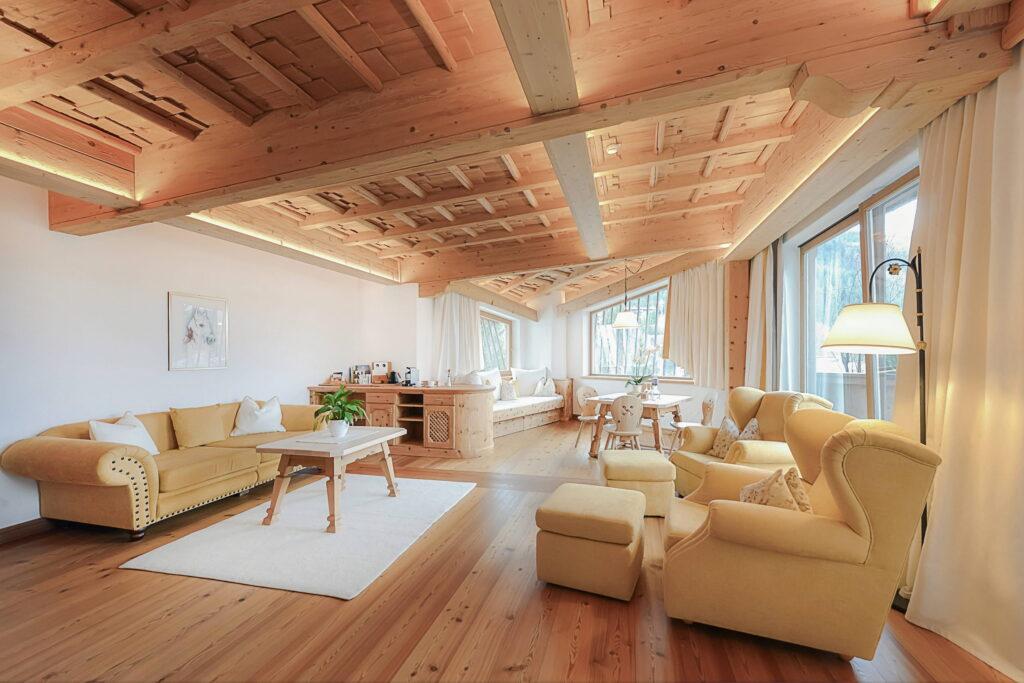 Ein geräumiger Raum mit Holzdecke und Holzböden, ausgestattet mit einem gelben Sofa und Sesseln. Große Fenster lassen Tageslicht herein und schaffen eine warme, gemütliche Atmosphäre.