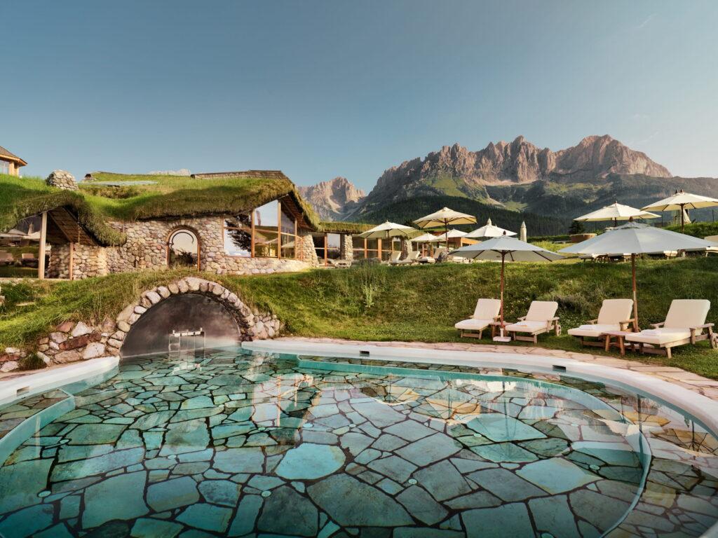 Outdoor pool at the Bioresort Stanglwirt in Tyrol, featuring a natural stone floor and stone arches, surrounded by lounge chairs with umbrellas in front of grass-covered buildings against the backdrop of the Wilder Kaiser mountains
