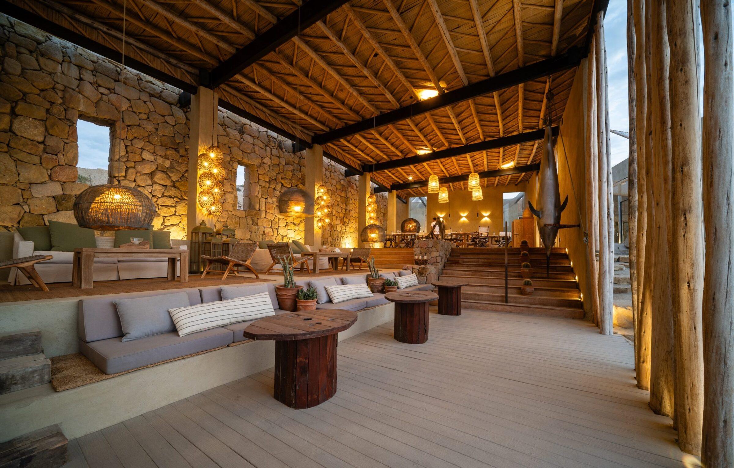 Geräumiger Loungebereich im Inkaterra Cabo Blanco mit Holzbalken, Steinwänden, eingebauten gepolsterten Sitzgelegenheiten, runden Holztischen und hängenden geflochtenen Lampen