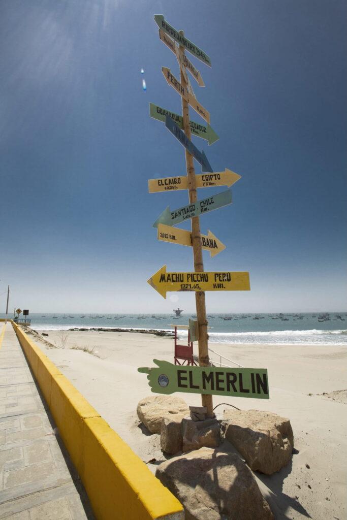 Holzschild mit mehreren Wegweisern, darunter „Machu Picchu“, an einem Sandstrand mit Meer und Booten im Hintergrund