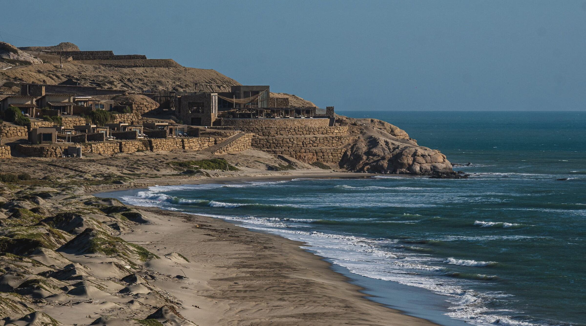 Die Gebäude des Küstenresorts Inkaterra Cabo Blanco auf felsigen Klippen mit Blick auf einen Sandstrand und das Meer unter strahlend blauem Himmel