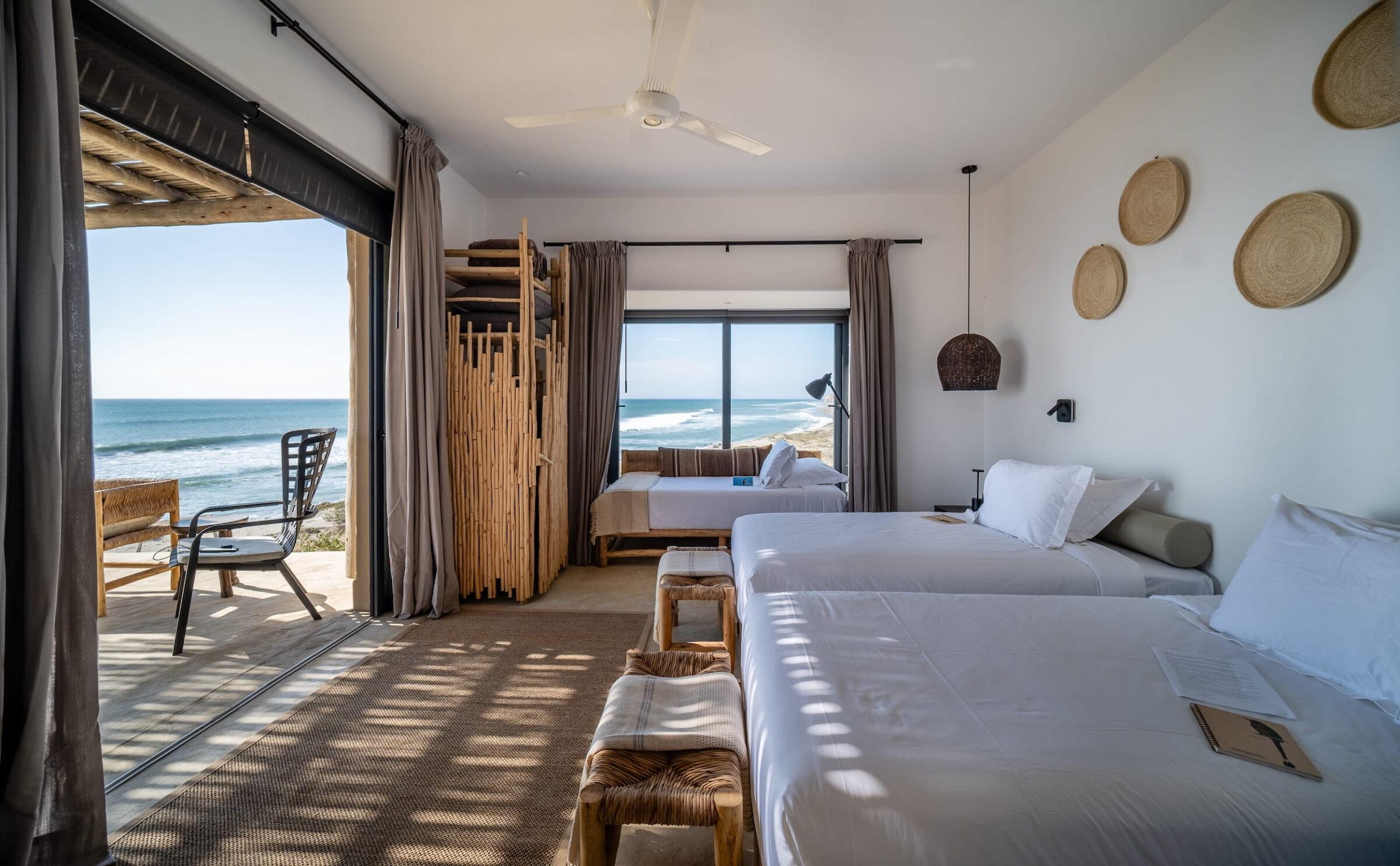 Hotelzimmer mit zwei Betten, Korbstühlen, an der Wand befestigten geflochtenen Körben und Glasschiebetüren, die auf eine Terrasse mit Stühlen und Blick auf das Meer im Inkaterra Cabo Blanco in Peru führen