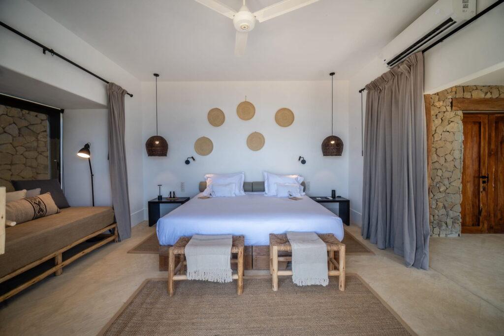 Minimalistisches Schlafzimmer im Inkaterra Cabo Blanco mit einem großen Bett, zwei Hockern mit geflochtenen Bezügen am Fußende, Pendelleuchten, runden Wanddekorationen und einem Türdurchgang mit Steinwand und Vorhängen