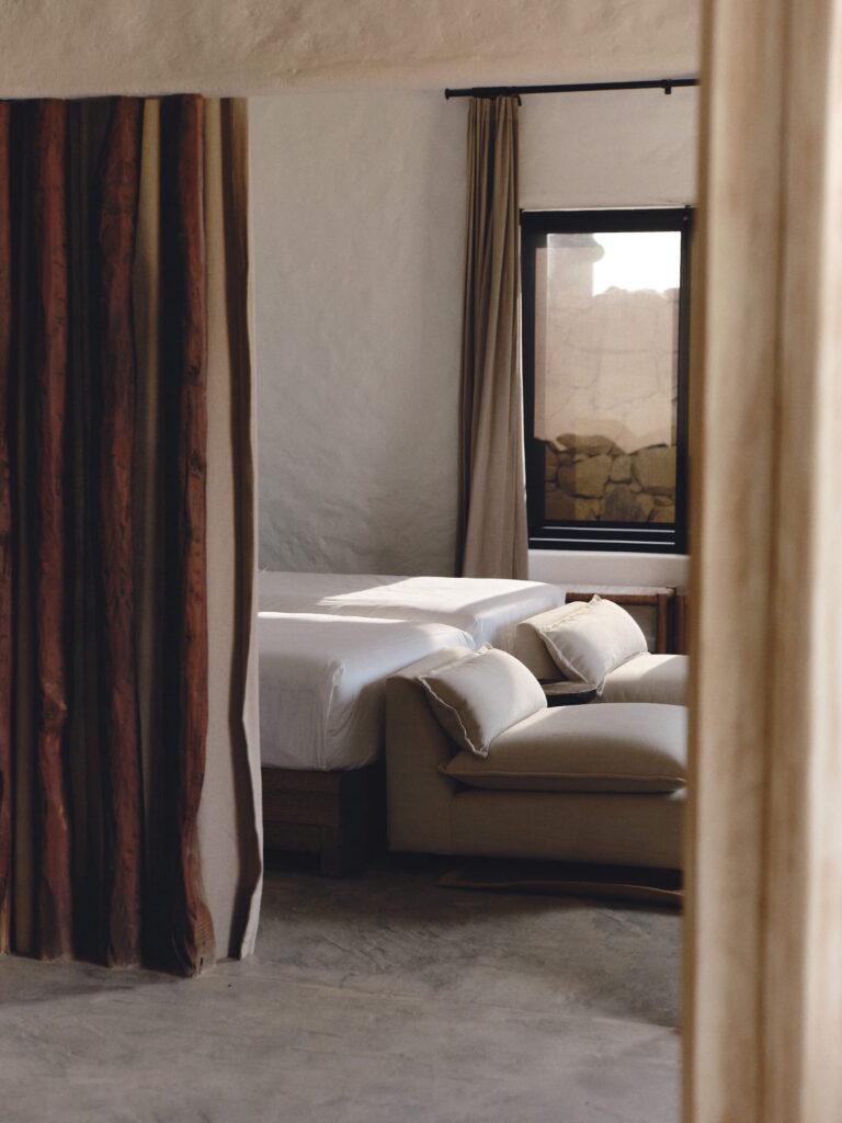 Blick in einen Raum mit einem Bett, einem Sofa mit Kissen, einem Fenster mit Vorhängen und einem gestreiften Stoff, der links hängt