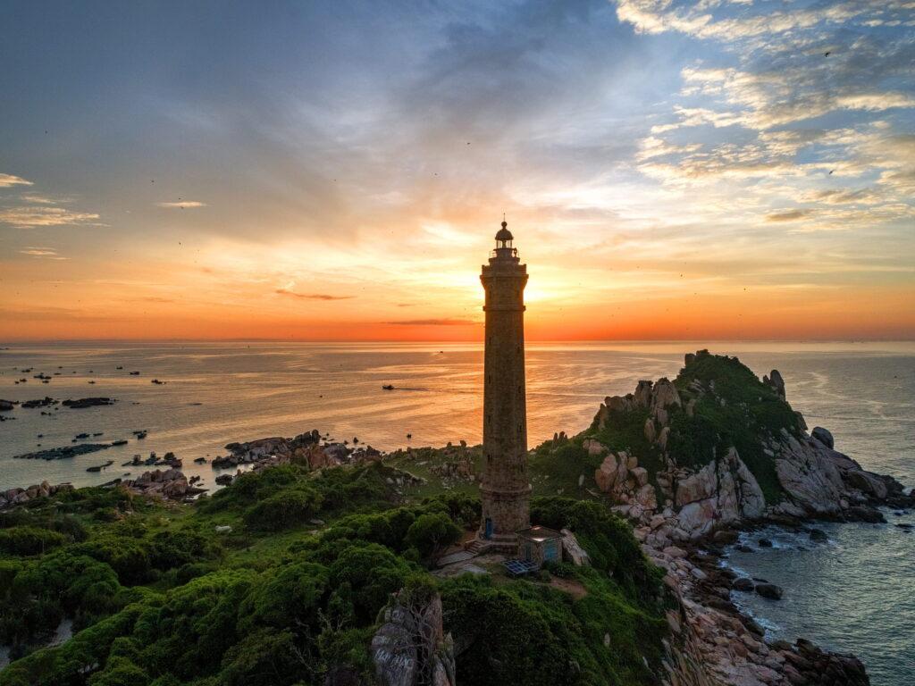 Der Leuchtturm von Ke Ga an einer felsigen, grün bewachsenen Küste in Vietnam bei Sonnenuntergang, mit vereinzelten Booten auf dem Meer