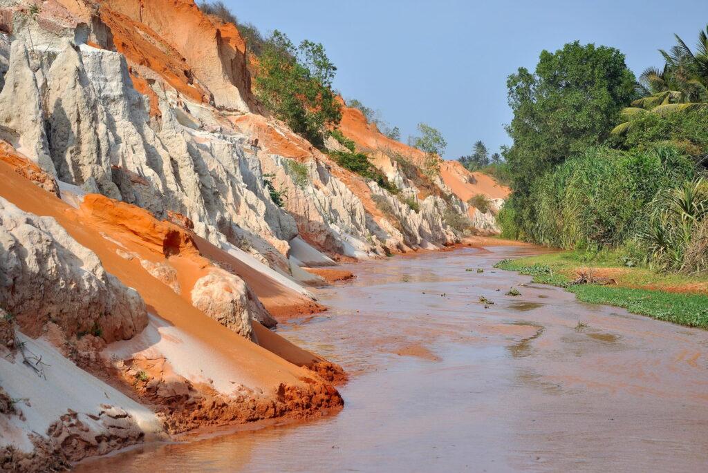 Der berühmte Fairy Stream in Vietnam fließt entlang erodierter orangefarbener und weißer Sandfelsen, rechts davon grüne Vegetation