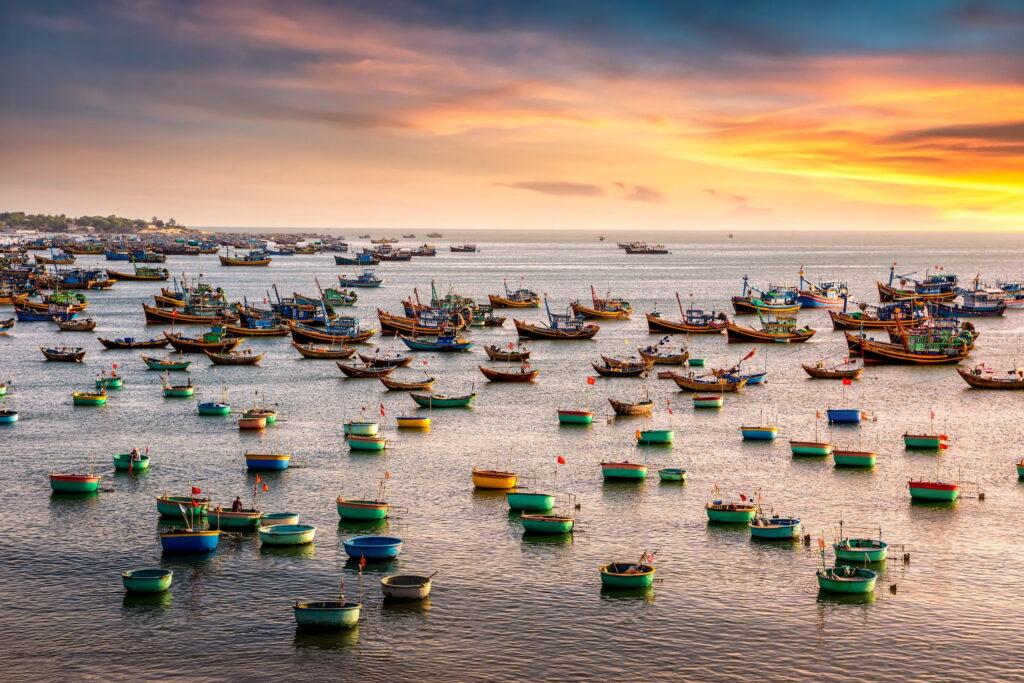Eine Gruppe von Fischerbooten und runden Korbbooten, die bei Sonnenuntergang in der Nähe von Mui Ne, Vietnam, auf dem ruhigen Meer treiben