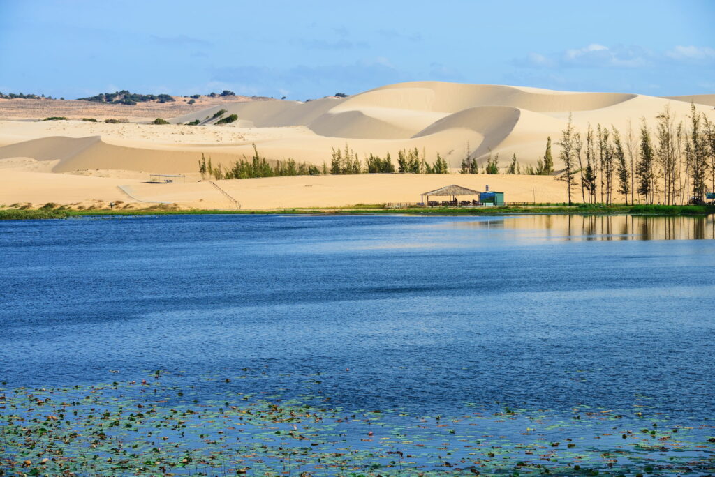 Ein großes Gewässer vor weißen Sanddünen mit spärlicher Vegetation und einem kleinen Unterstand in Ufernähe bei Mui Ne in Vietnam