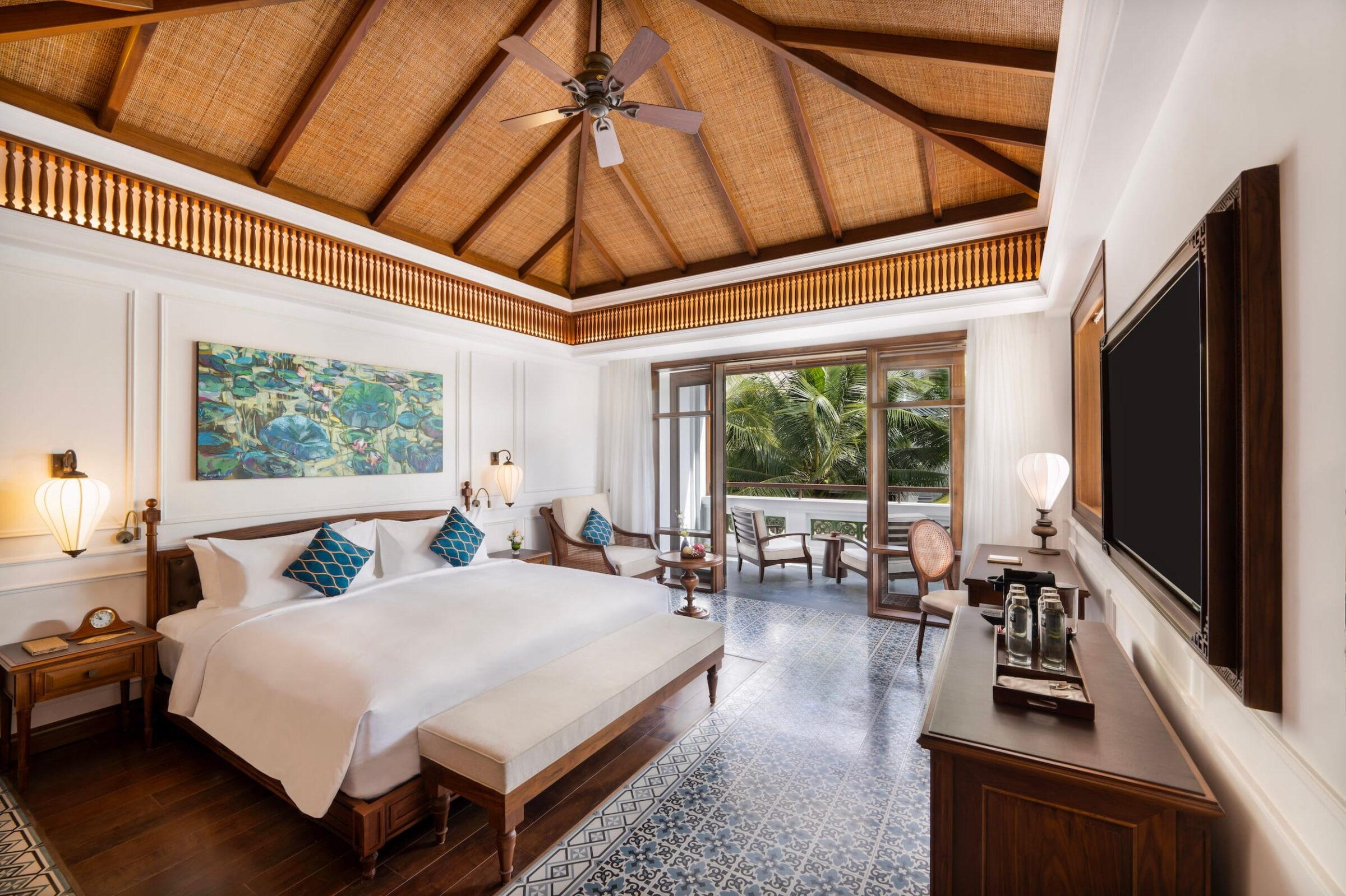 Geräumiges Hotelzimmer im luxuriösen Resort „The Anam Mui Ne“ mit einem großen Bett, gemusterten Bodenfliesen, Holzmöbeln, einem Deckenventilator und einem Balkon mit Sitzgelegenheiten im Freien und Blick ins Grüne
