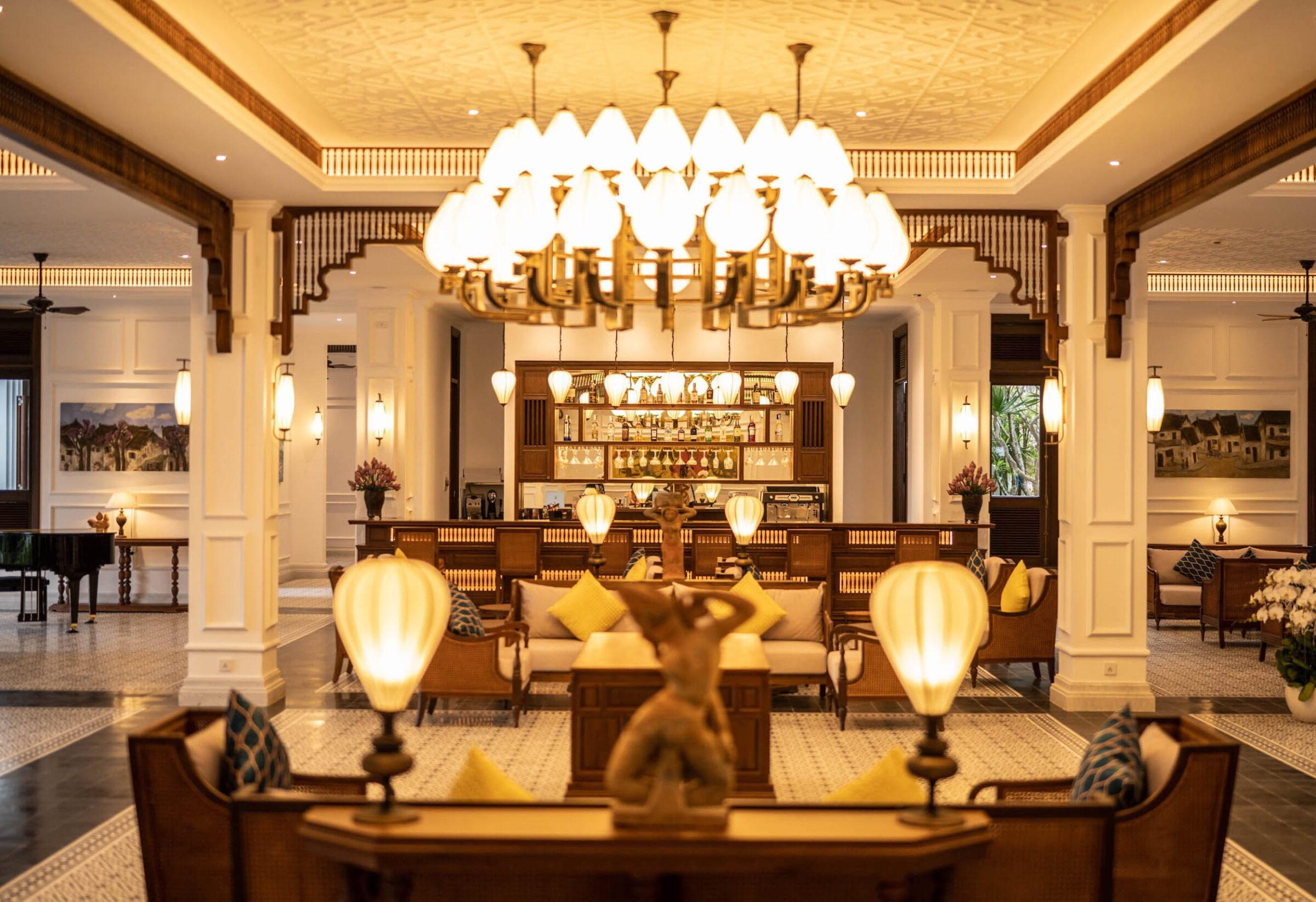 Die Lobby des Resorts „The Anam Mui Ne“ in Vietnam mit Holzmöbeln, einem zentralen Kronleuchter, dekorativen Lampen und einer Statue auf einem Tisch