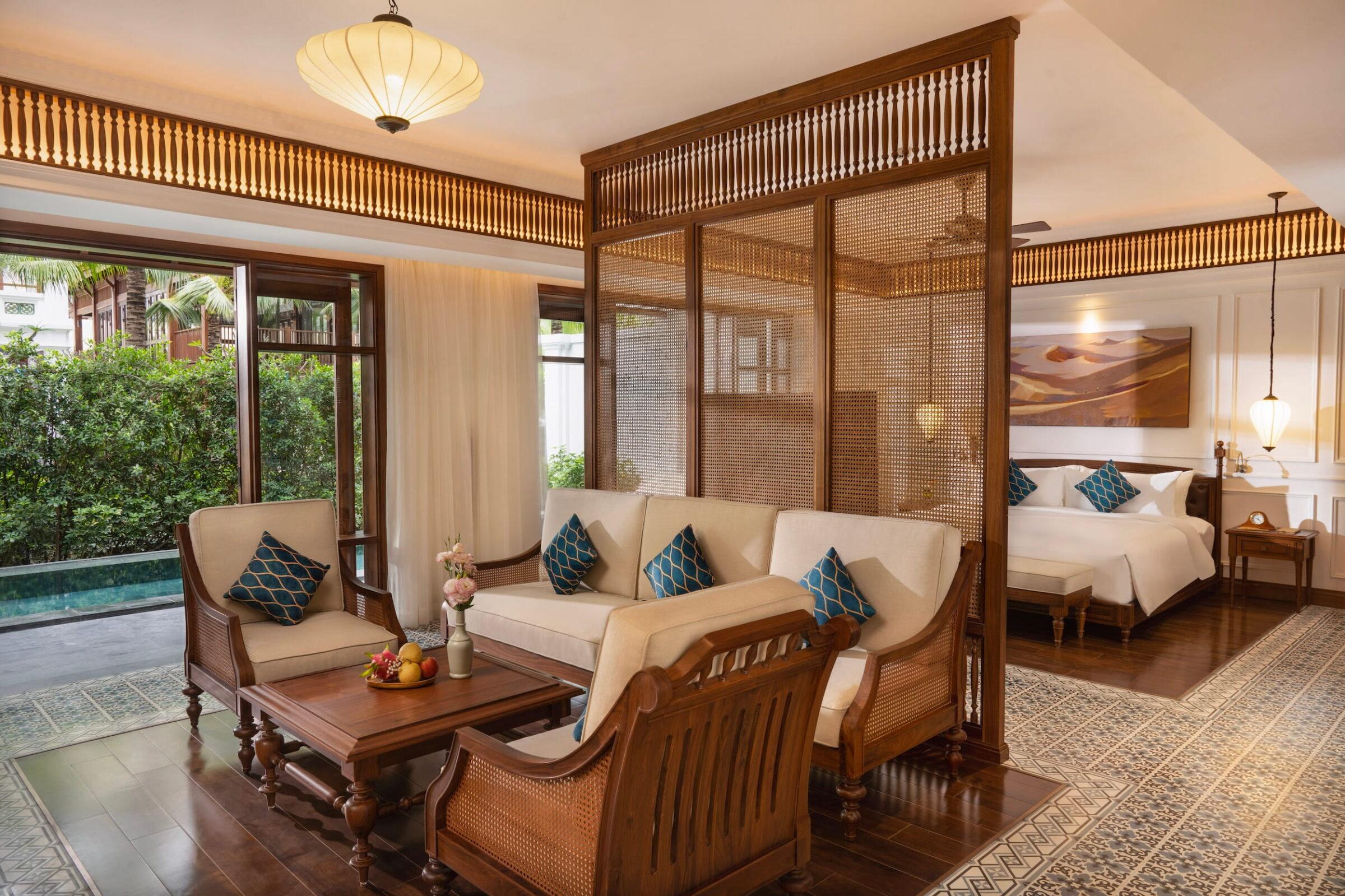 Wohnbereich mit Holzmöbeln und blau gemusterten Kissen, durch eine Trennwand aus Holzgitter von einem Schlafzimmer mit großem Bett und Nachttischlampen getrennt, im vietnamesischen Resort „The Anam Mui Ne“