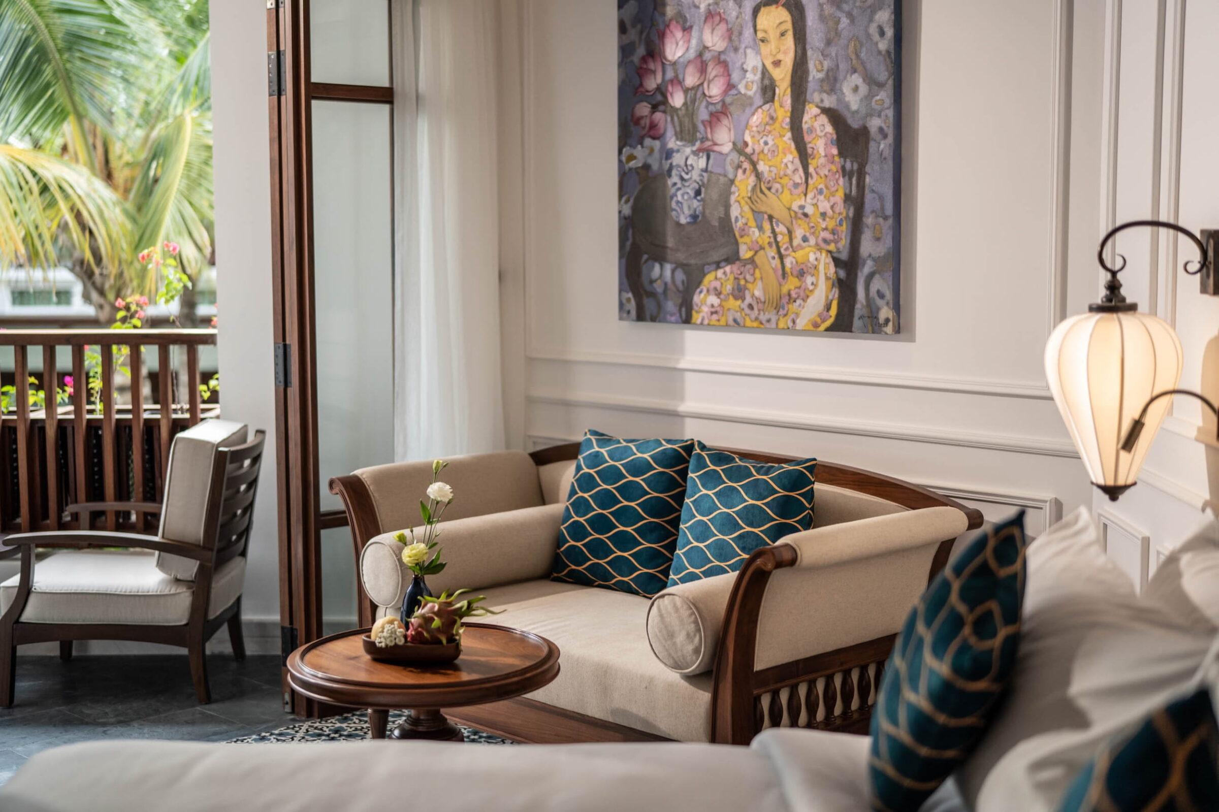 Sitzecke in einem Resort-Zimmer mit einem beigen Sofa, gemusterten blauen Kissen, einem runden Holztisch mit einer Blumenvase, einem Gemälde einer Frau an der Wand und einer Hängelampe