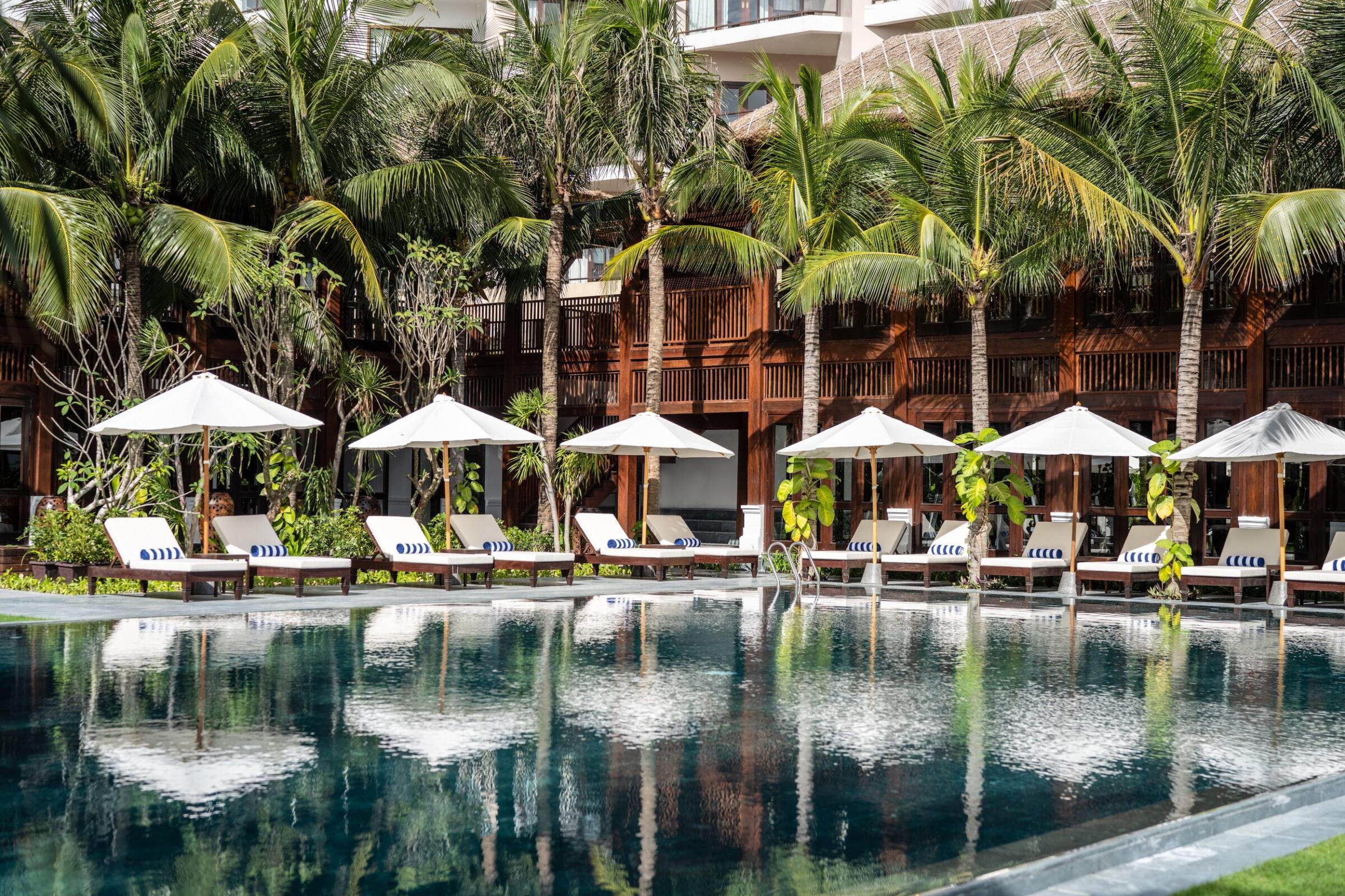 Poolbereich mit Liegestühlen und Sonnenschirmen vor Palmen und einem Gebäude im Resort „The Anam Mui Ne“ in Vietnam