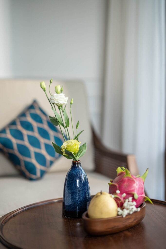 Eine blaue Keramikvase mit weißen und gelben Blumen auf einem runden Holztisch neben einer Holzschale, in der eine Drachenfrucht, eine Birne und kleine weiße Blumen liegen, im Hintergrund ein beiges Sofa und ein blau gemustertes Kissen