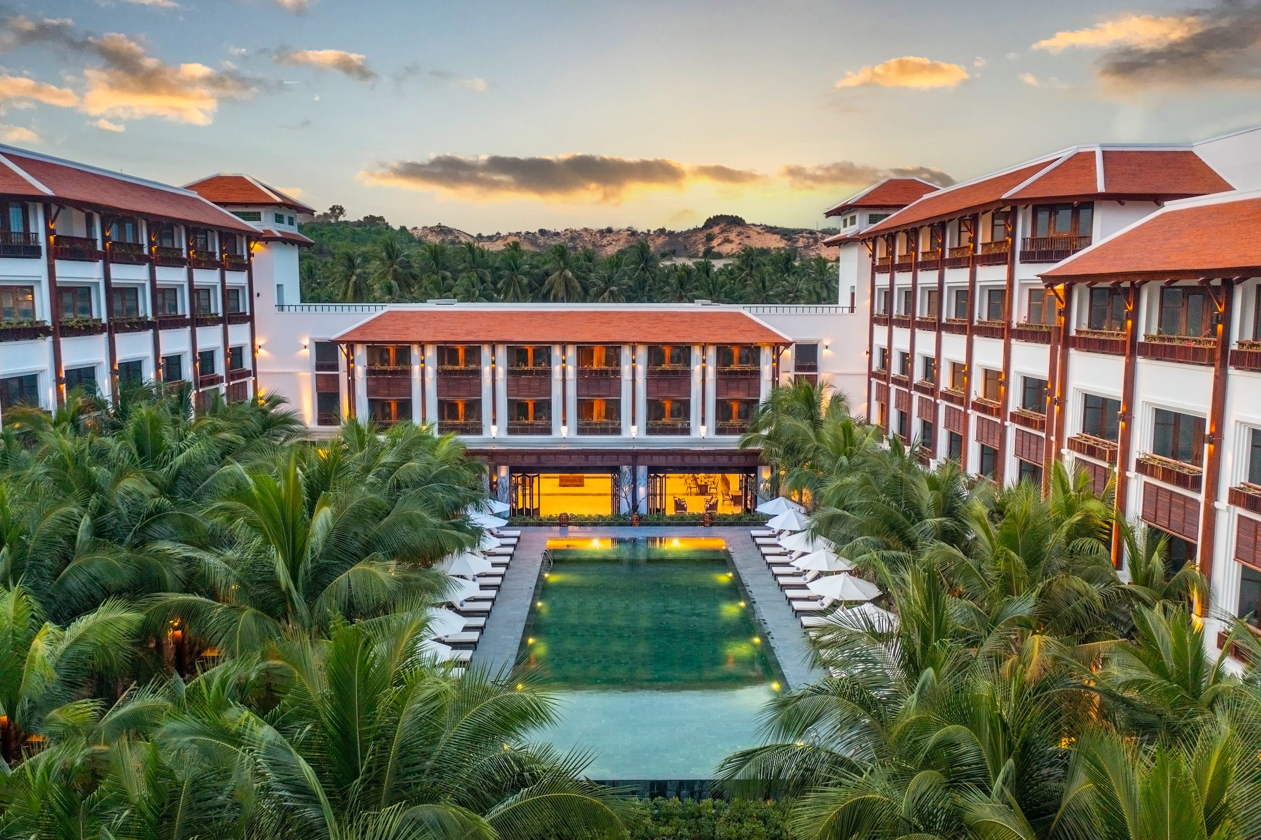 Symmetrische Ansicht des Anam Mui Ne Resorts in Vietnam mit einem zentralen, rechteckigen Pool, der von Palmen umgeben ist und von zwei vierstöckigen Gebäuden mit Balkonen flankiert wird – unter einem teilweise bewölkten Himmel bei Sonnenuntergang