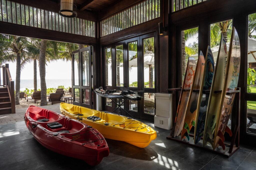 Zwei Kajaks, eines rot und eines gelb, stehen in einem Holzpavillon im „The Anam Mui Ne“, daneben stehen Surfbretter aufrecht, und draußen sind Palmen zu sehen