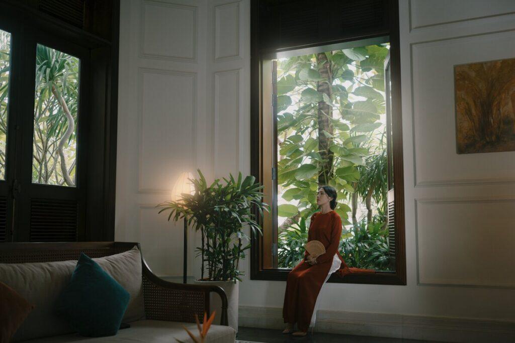 Eine Frau in einem roten, traditionellen Ao-Dai-Kleid sitzt auf einem breiten Fensterbrett; draußen sind grüne Pflanzen zu sehen, in einem sanft beleuchteten Raum im „The Anam Mui Ne“ in Vietnam