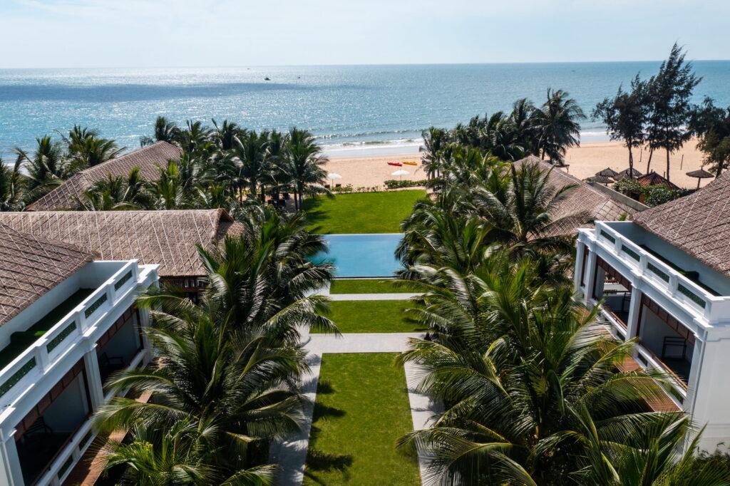 Blick auf das luxuriöse „The Anam Mui Ne“ in Vietnam mit zwei Gebäudereihen und Palmen, die eine zentrale Rasenfläche säumen, die zu einem rechteckigen Pool und einem Sandstrand mit dem Meer im Hintergrund führt