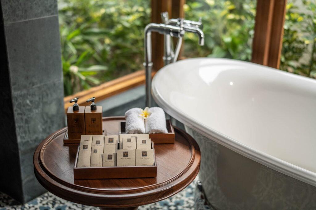 Runder Holztisch mit Toilettenartikeln und aufgerollten Handtüchern neben einer freistehenden Badewanne mit Chromarmatur in einer luxuriösen Suite im „The Anam Mui Ne“ in Vietnam