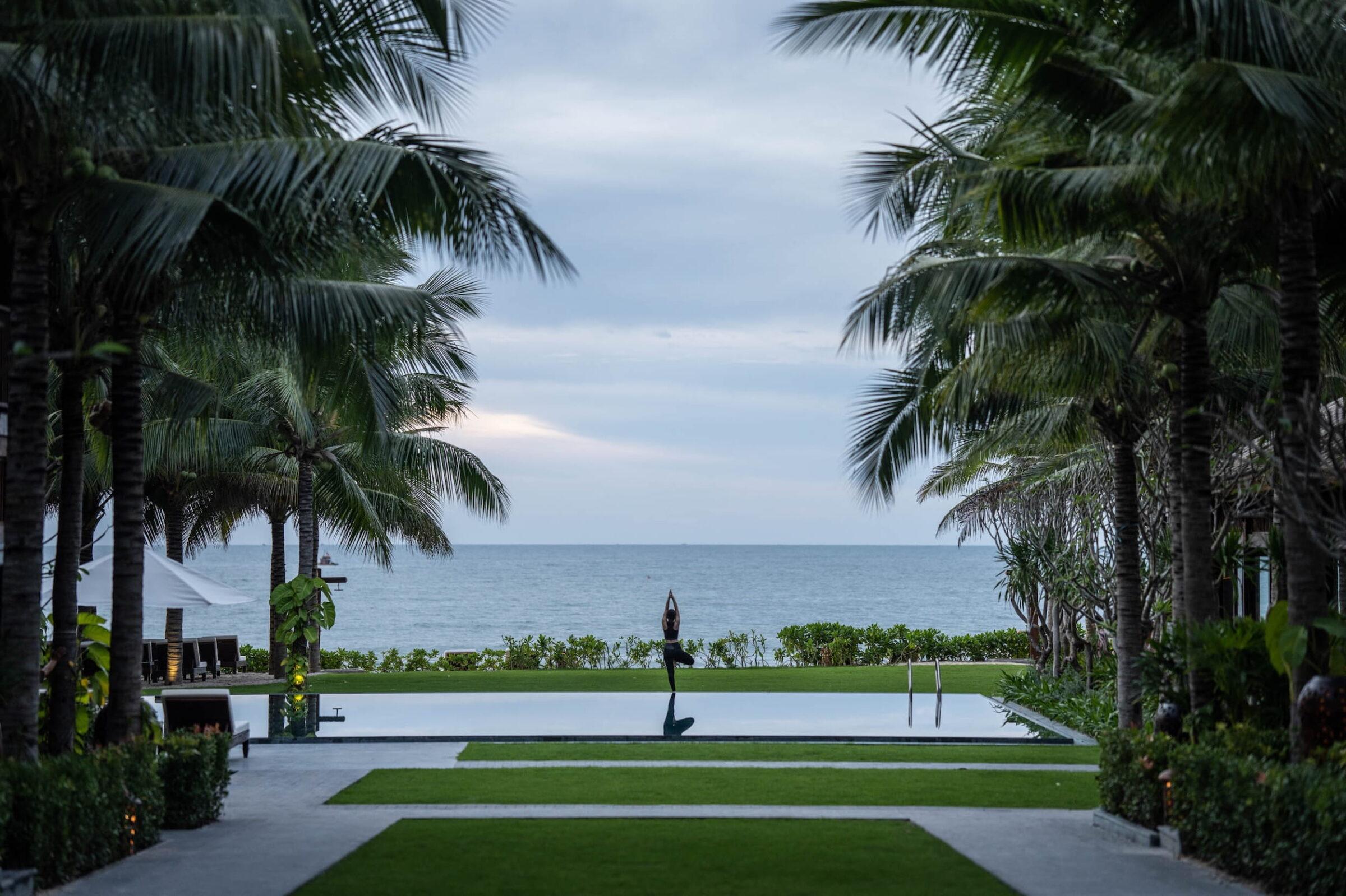 Eine Person, die in einer Yoga-Pose auf einer Plattform am Pool steht, während Palmen den Blick auf das Meer im Resort „The Anam Mui Ne“ in Vietnam einrahmen