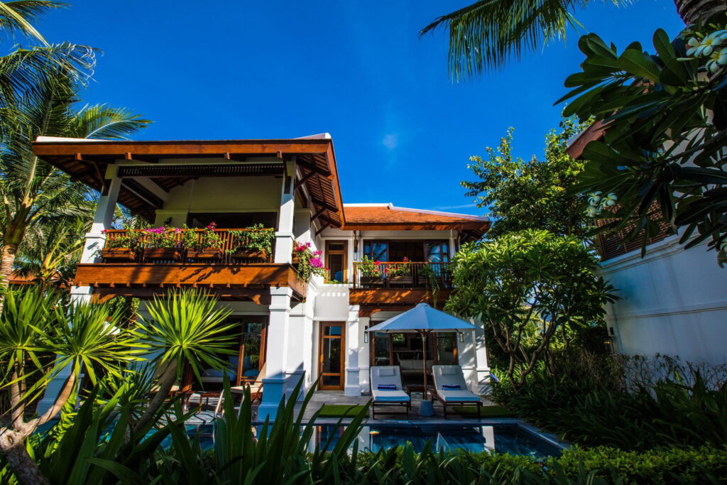 Zweigeschossige Villa mit Holzbalkon, Liegestühlen am Pool unter einem Sonnendach, umgeben von tropischen Pflanzen