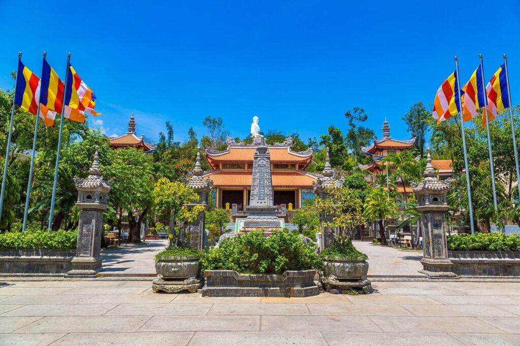 Vorderansicht der Long-Son-Pagode mit einem zentralen Steinmonument, einer weißen Buddha-Statue dahinter, flankiert von traditionellen vietnamesischen Tempelgebäuden und Reihen bunter buddhistischer Fahnen