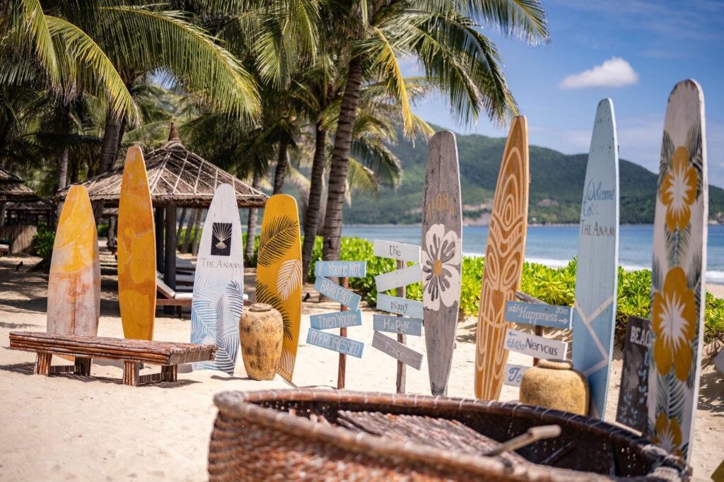 Im Resort „The Anam Cam Ranh“ in Vietnam stehen mehrere bunte Surfbretter und Holzschilder mit Aufschriften im Sand vor Palmen und mit Blick aufs Meer