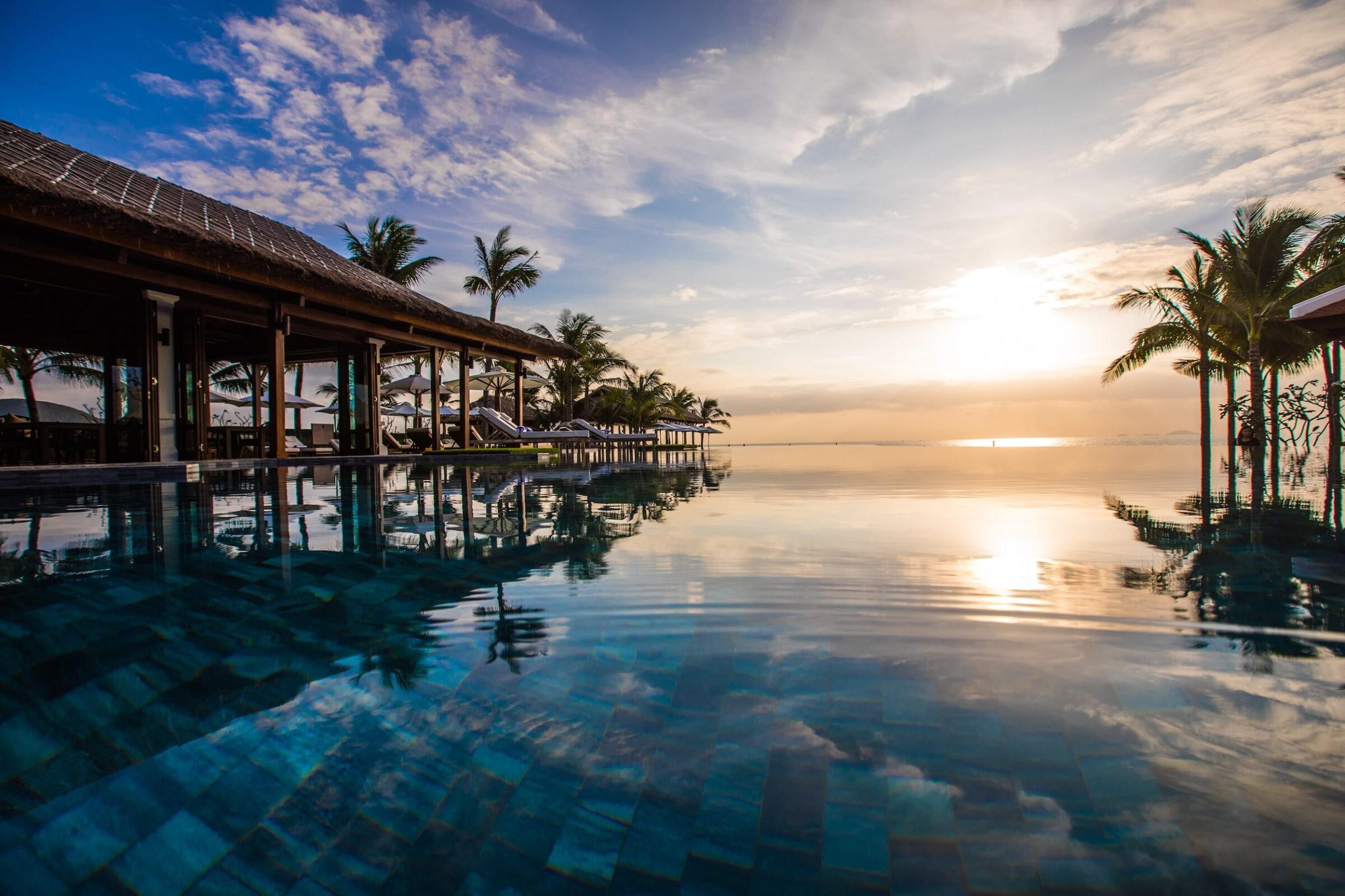 Ein großer Infinity-Pool mit ruhiger Wasseroberfläche spiegelt den Himmel bei Sonnenuntergang wider. Im Hintergrund sind Palmen und ein langes, offenes Gebäude mit einem traditionellen vietnamesischen Dach zu sehen. Liegestühle mit weißen Kissen säumen den Rand des Pools im The Anam Cam Ranh.