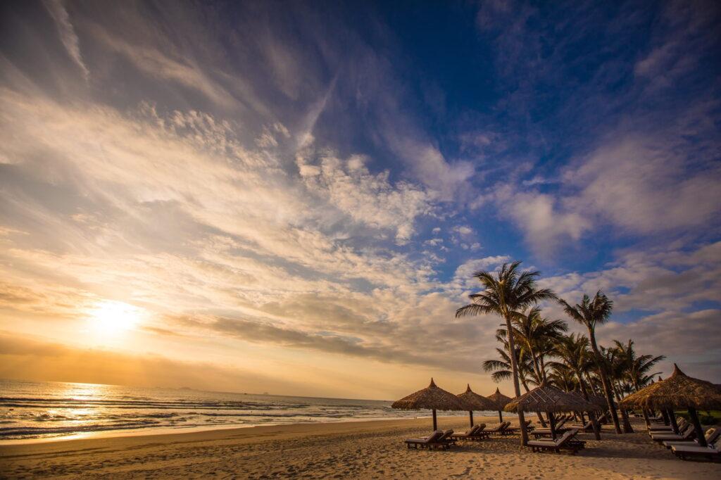Der Strand an der Cam-Ranh-Bucht bei Sonnenaufgang, mit Palmen und strohgedeckten Sonnenschirmen am Ufer