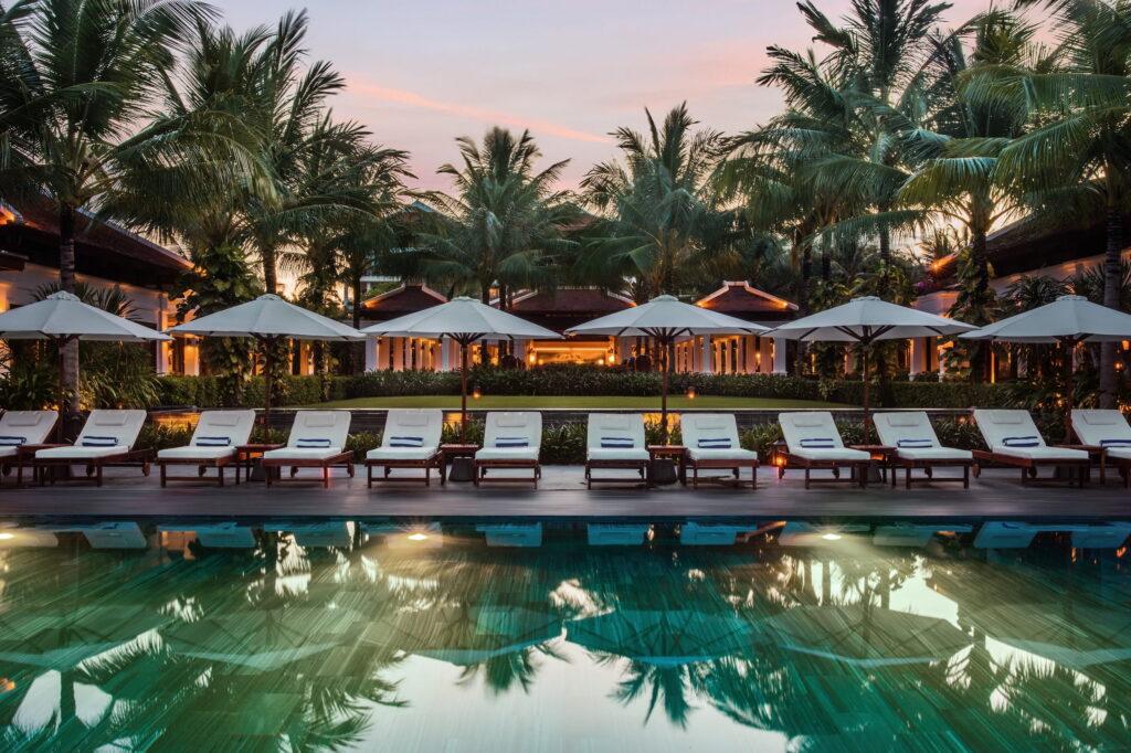 Pool mit Liegestühlen und Sonnenschirmen vor tropischen Bäumen und Resortgebäuden in der Abenddämmerung