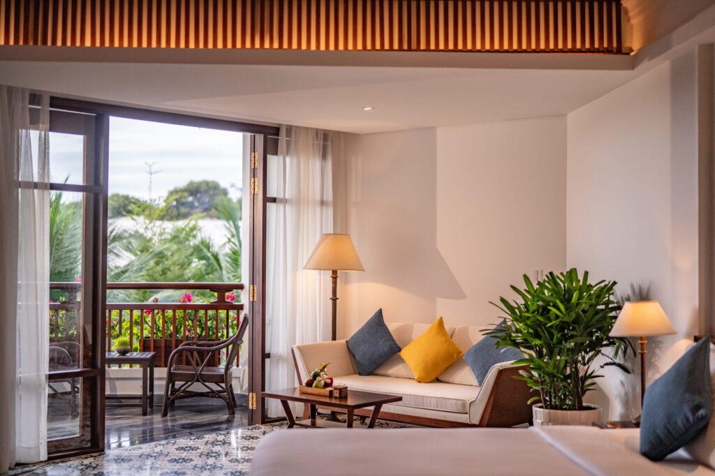 Hotelzimmer im „The Anam Cam Ranh“ Resort mit einem Bett, einem Sofa mit Kissen, zwei Lampen, einem Couchtisch, Topfpflanzen und einer offenen Tür, die auf einen Balkon mit Stühlen und viel Grün im Freien führt
