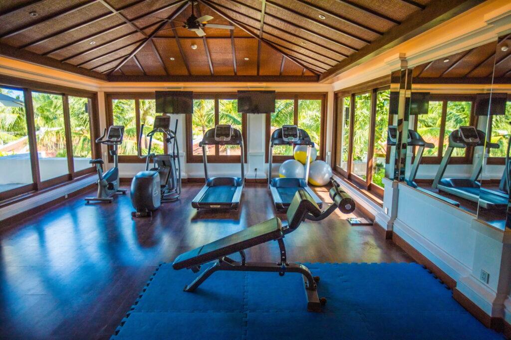 Fitnessraum mit Heimtrainern, Laufbändern, einer Hantelbank auf blauen Matten und großen Fenstern mit Blick auf tropische Vegetation
