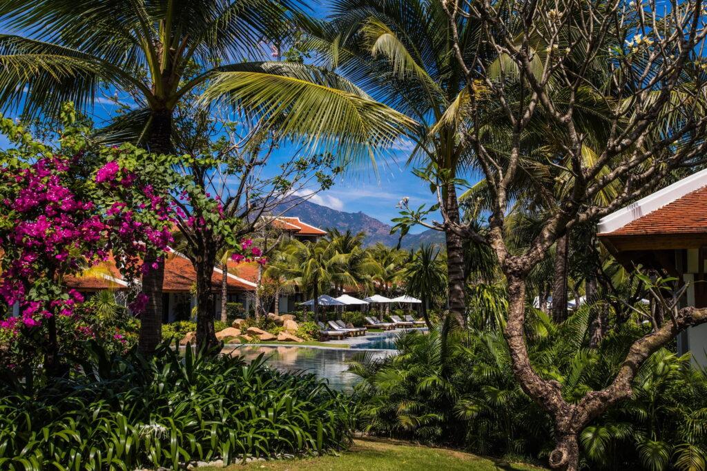 Eine Resort-Lagune, umgeben von Palmen, blühenden Sträuchern, Sonnenliegen mit Sonnenschirmen und Gebäuden mit rotgedeckten Dächern unter blauem Himmel