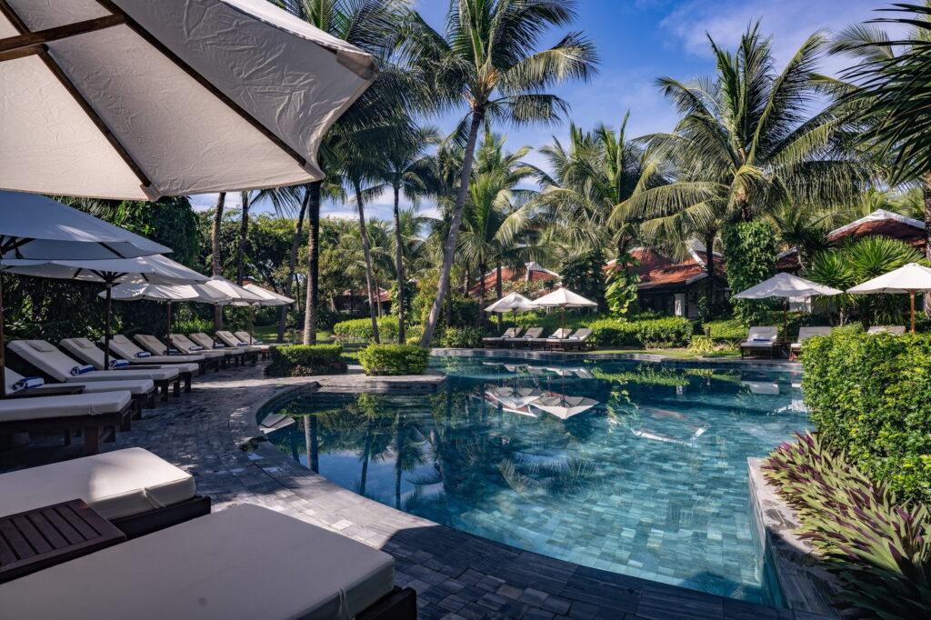 Der Poolbereich des Resorts „The Anam Cam Ranh“ mit Liegestühlen unter Sonnenschirmen und Palmen, die einen gefliesten Pool im Lagunenstil umgeben