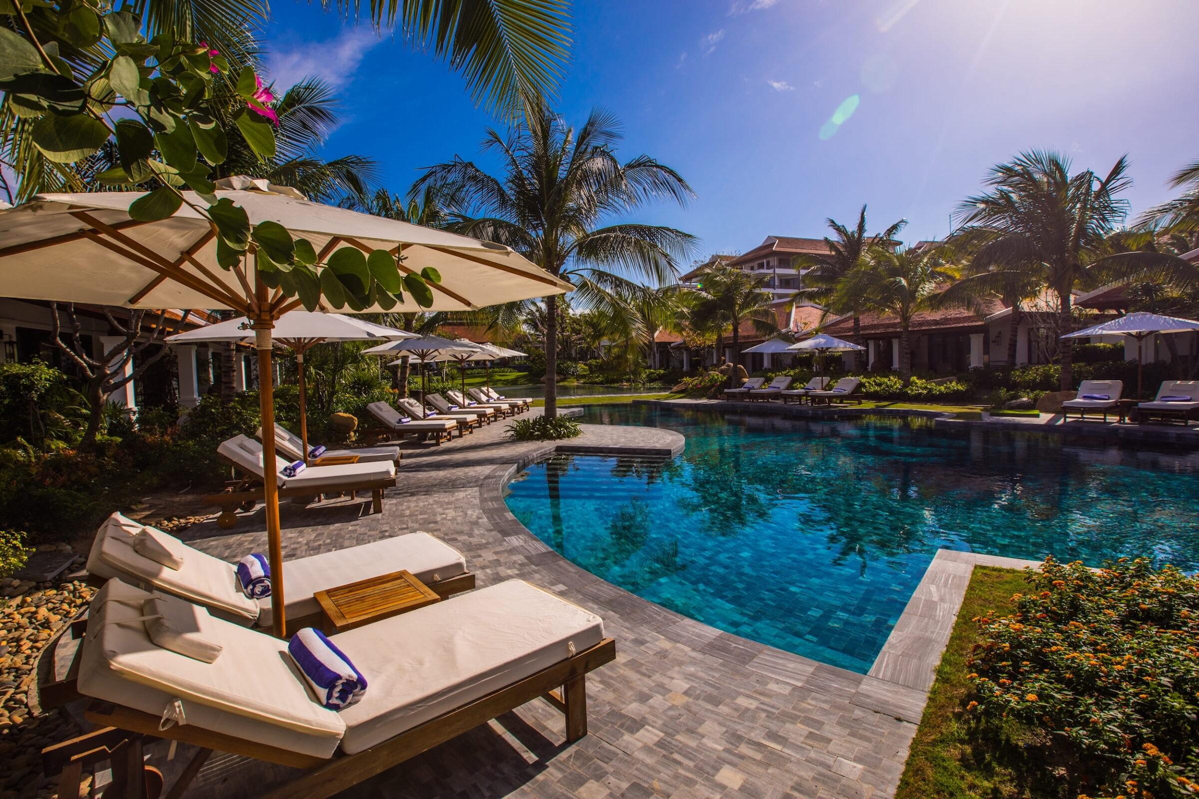 Der Pool im luxuriösen „The Anam Cam Ranh“ mit Liegestühlen und Sonnenschirmen, umgeben von Palmen und Gebäuden unter strahlend blauem Himmel